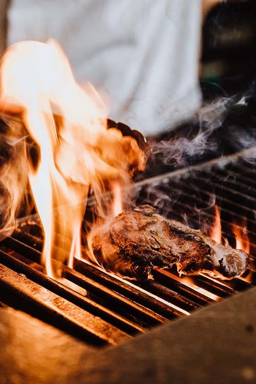 Steak on the grill with flames. Chef holding tongs over the steak.