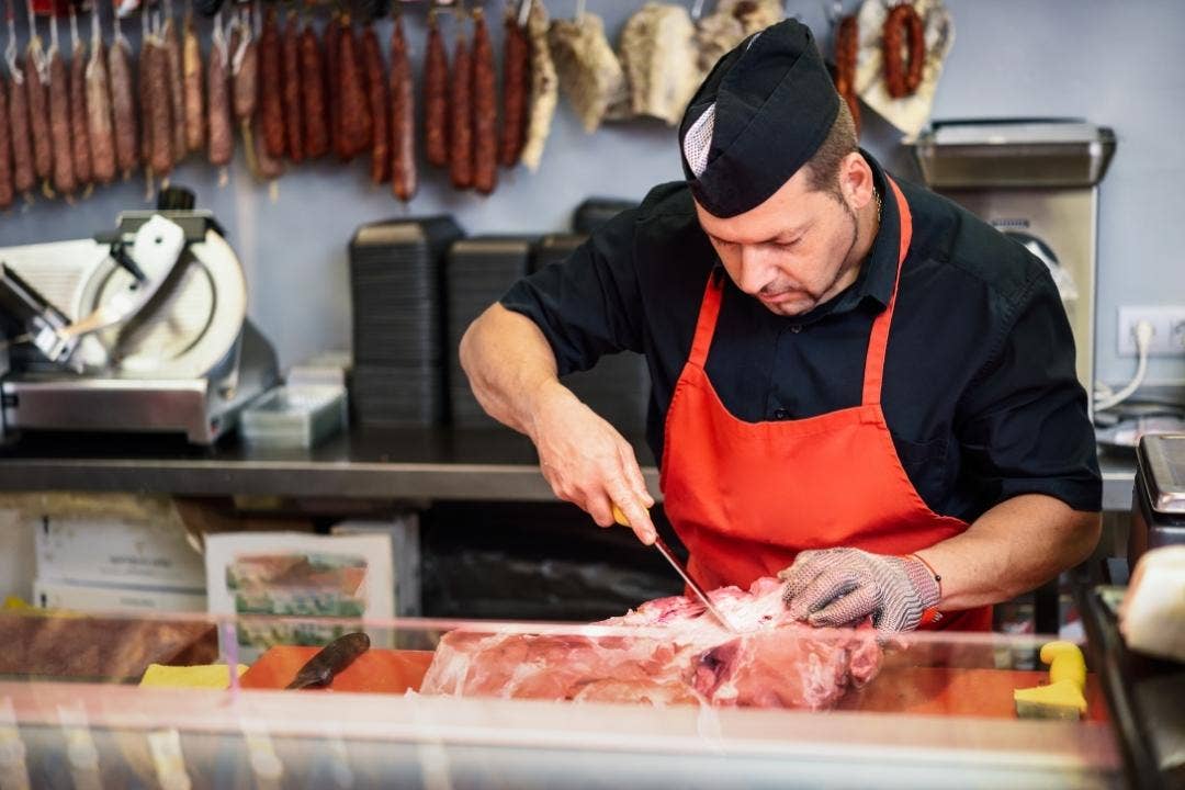 Butcher trimming fresh meat behind counter with sausages hanging in background