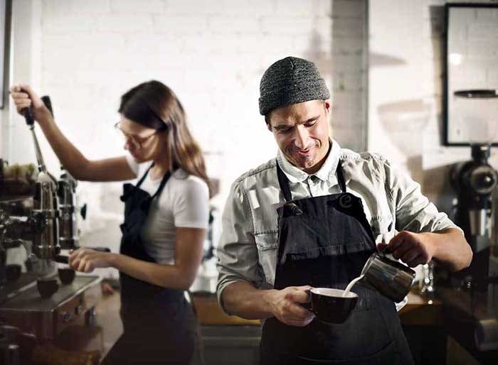 A male barista pours milk into a coffee cup while a female barista uses an espresso machine in the background.
