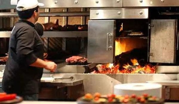 Chef operating a wood-fired oven with steaks and flames in a restaurant kitchen.