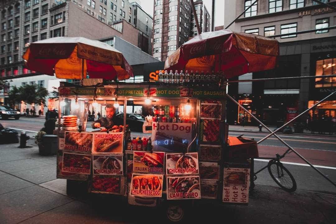 Hot dog vendor preparing food at a colorful street cart in a city setting