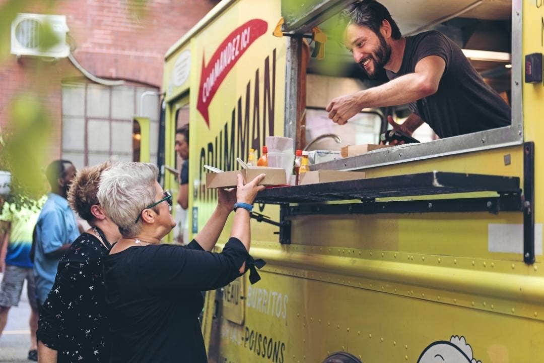 Man serving food from a yellow food truck to smiling customers outdoors