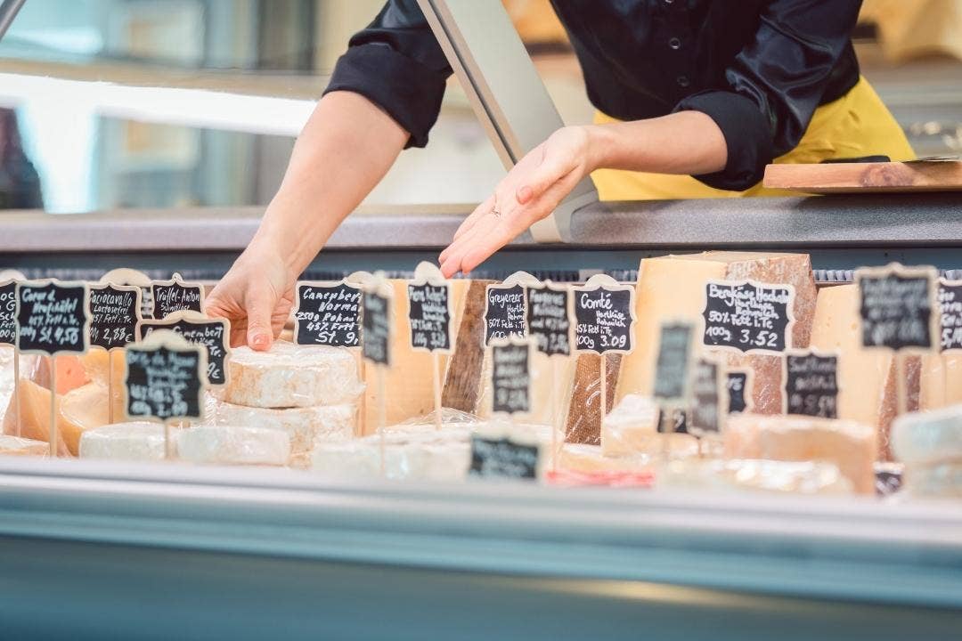 Deli worker arranging labeled gourmet cheeses inside a refrigerated display case.