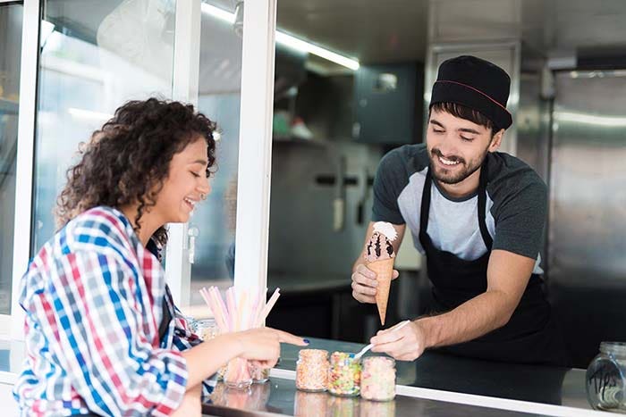 A beautiful girl in a plaid shirt ordered chocolate ice cream. A man in a mobile diner gives her ice cream. They both smile at each other. The man adds to the creamy whipped cream