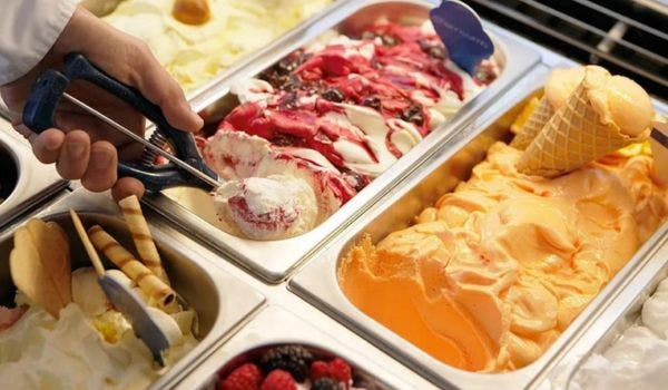 Hand scooping various flavors of gelato and toppings from an ice cream display case.