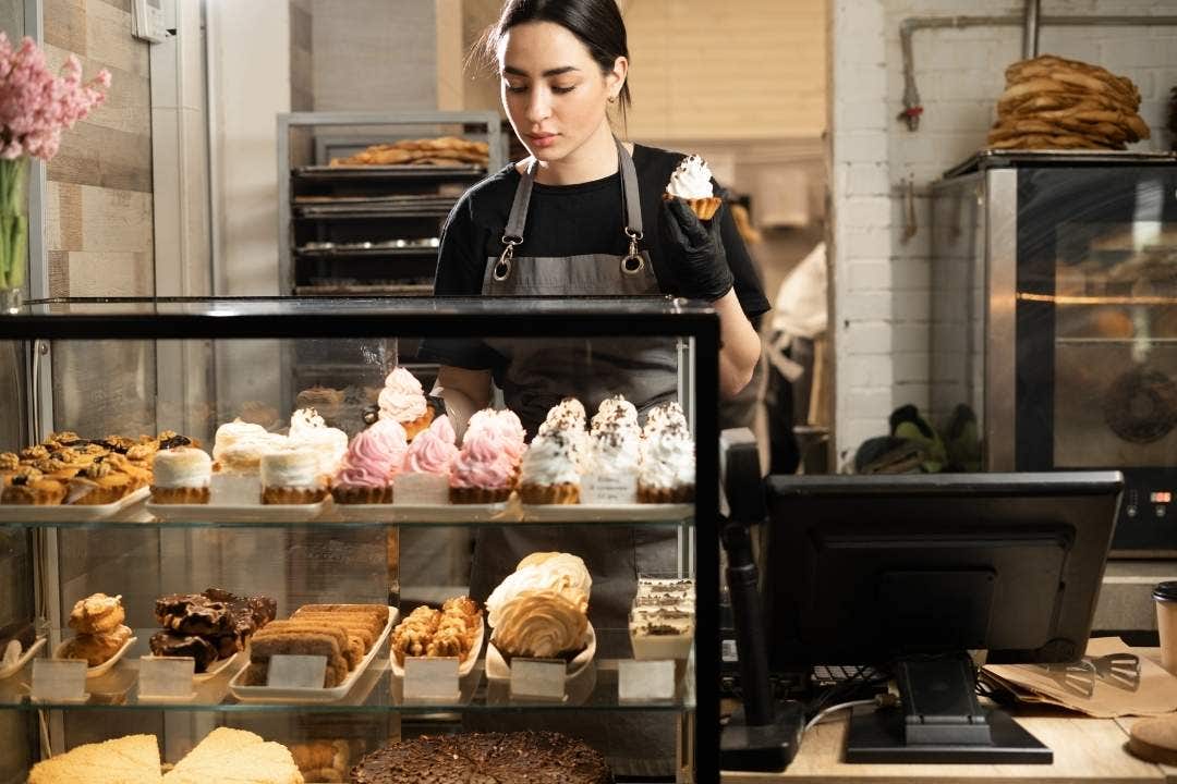 Bakery worker behind pastry display filled with cupcakes, cookies, and desserts.