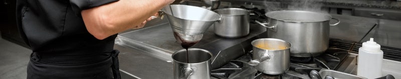Chef pouring sauce through funnel into pot on busy commercial kitchen stovetop