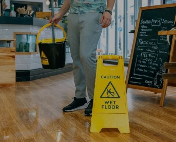 Person walking on wood floor with a mop bucket and mop in one hand as they walk by a yellow sign that says "Caution Wet Floor."