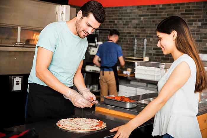 Chef wearing gloves preparing pizza adding pepperoni while customer looking at pizza in pizzeria