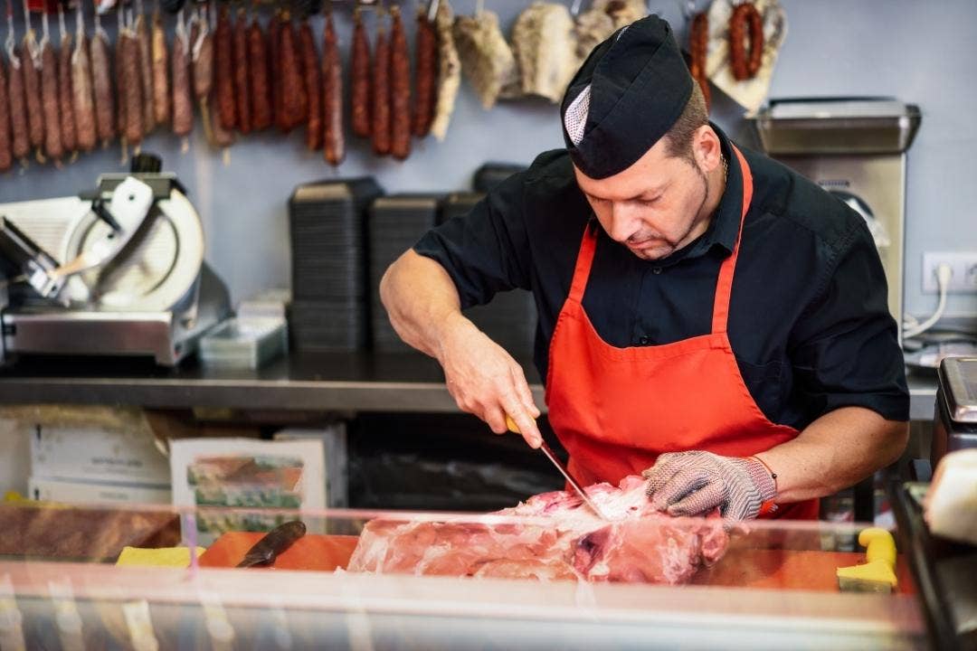 Butcher cutting fresh meat beside hanging sausages and meat slicer