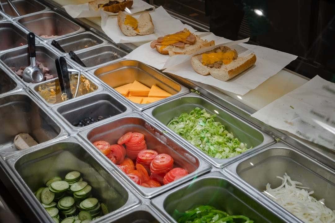 Sub sandwich prep station with sliced vegetables, cheese, and sandwich buns
