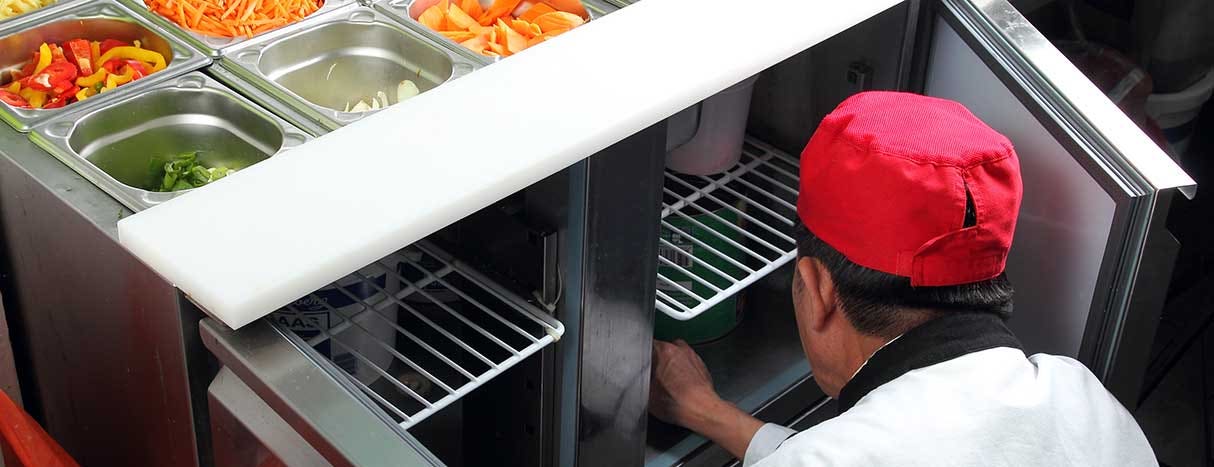 A cook in a kitchen kneeling to look into the bottom refrigerator of a mega top prep table.
