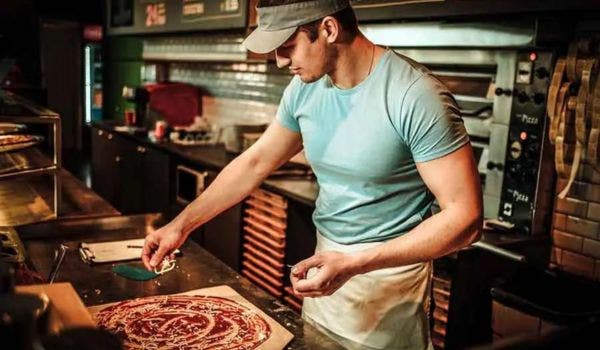 Male chef in a pizza kitchen adding toppings to a freshly prepared pizza.