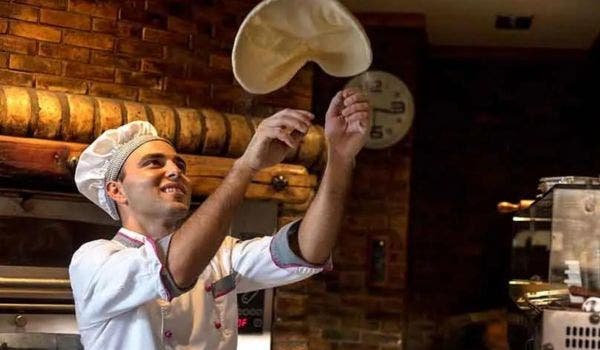 Pizza chef tossing dough in the air in a brick-oven restaurant setting.
