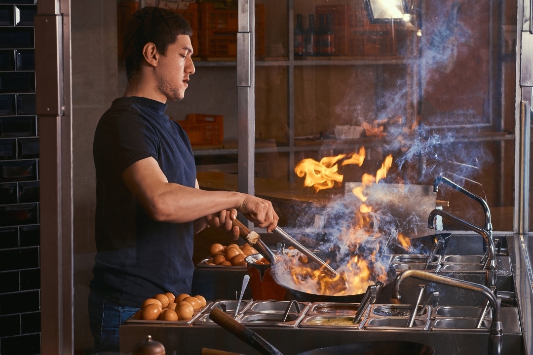 Chef cooking with high flames on wok range in a busy restaurant kitchen.