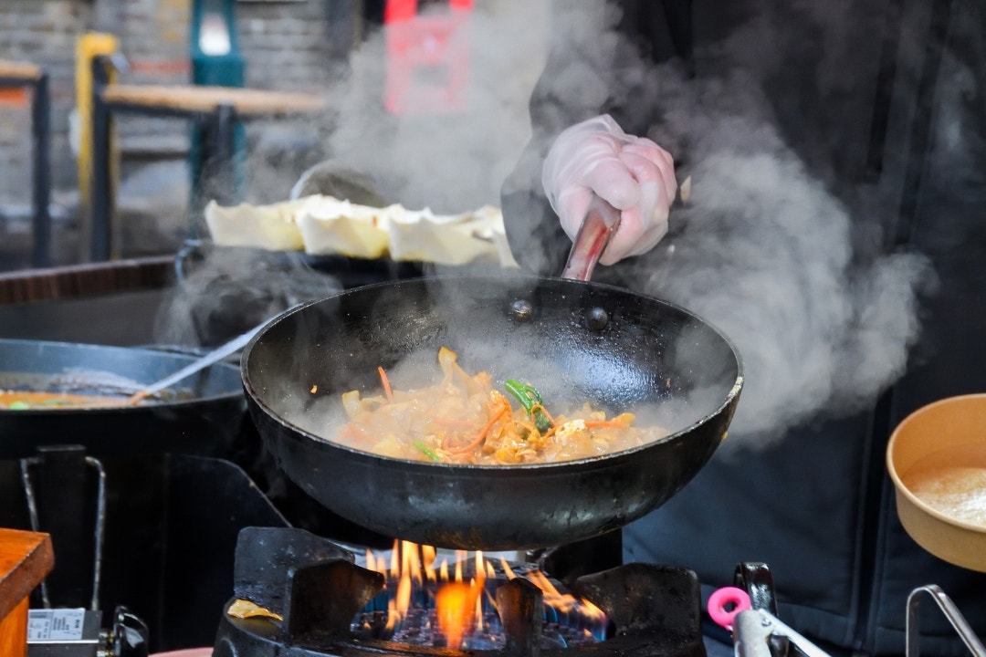 Chef stir-frying vegetables in wok on commercial gas range at outdoor market.