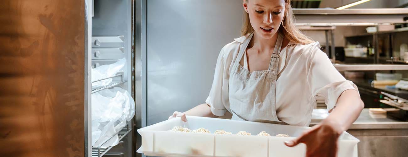 Young blonde woman wearing apron putting food in refrigerator while working in restaurant kitchen