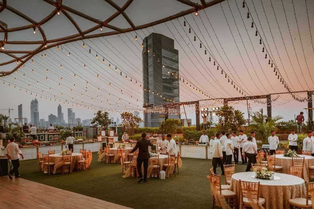 Outdoor banquet event setup with string lights and servers under a city skyline