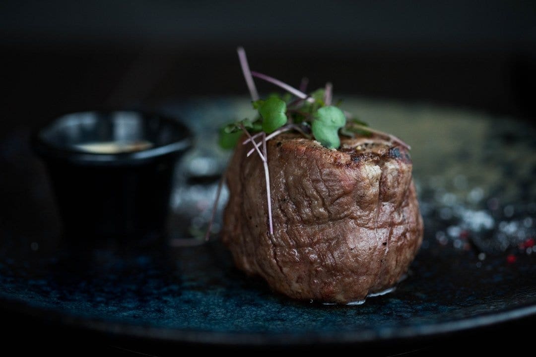 Grilled filet mignon topped with microgreens on a dark plate with sauce cup