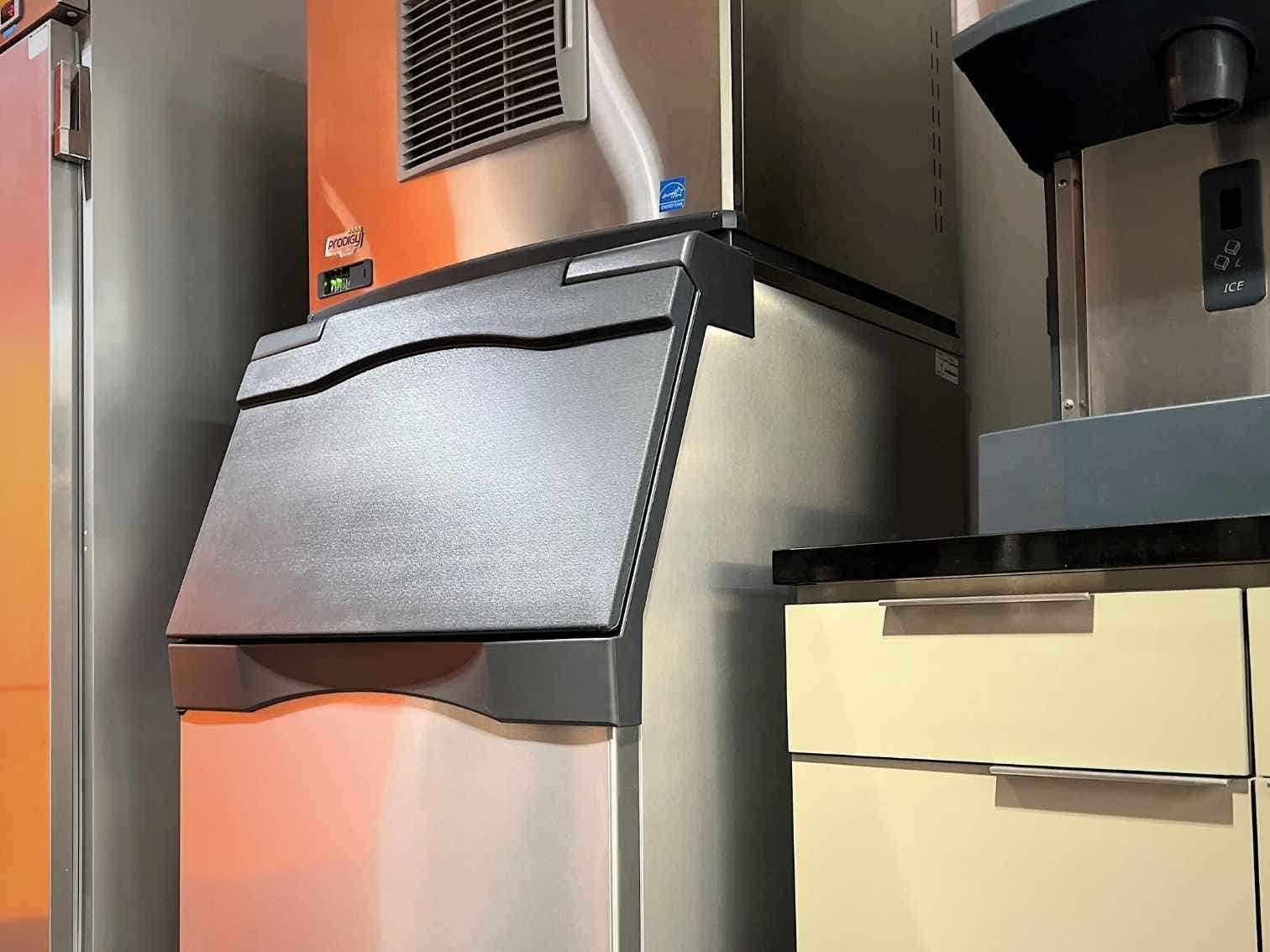 An stainless steel ice machine and bin with a black door situated between a stainless steel refrigerator and a cabinet with harvest gold doors and drawers.