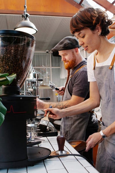 Male and female barista cleaning bean grinder and preparing orders.