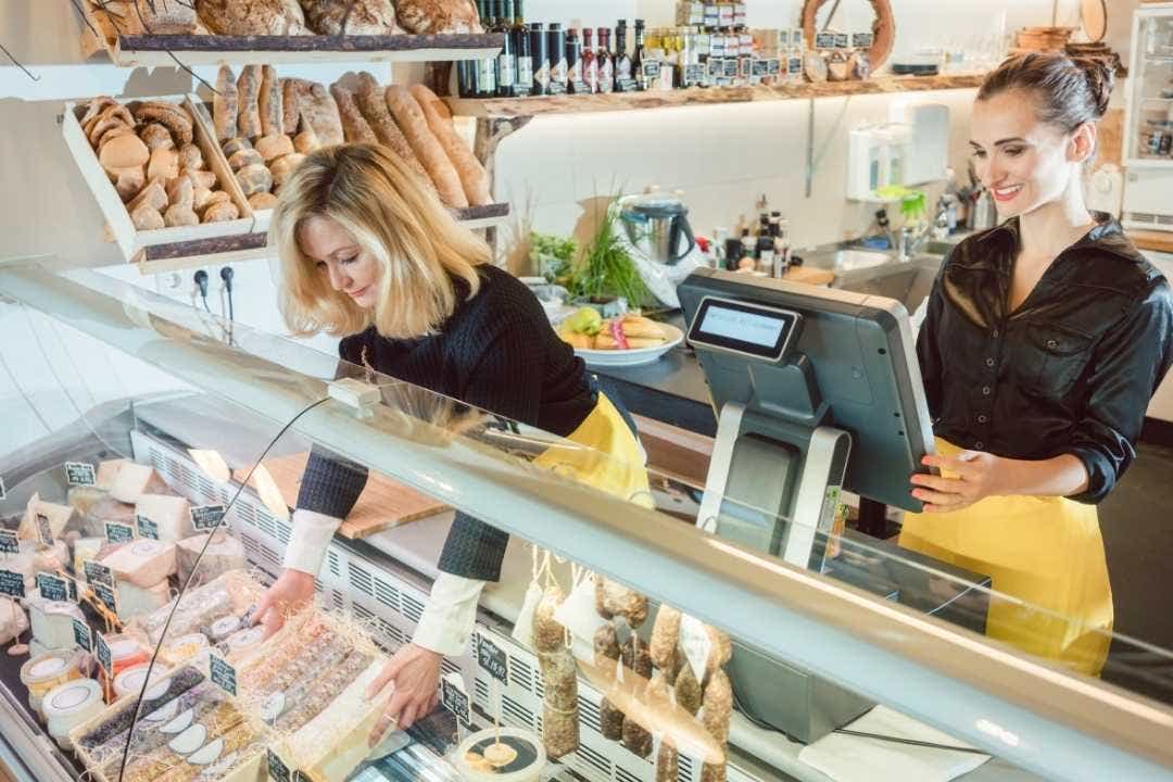 Two women working behind a deli counter with cheeses and baked goods on display