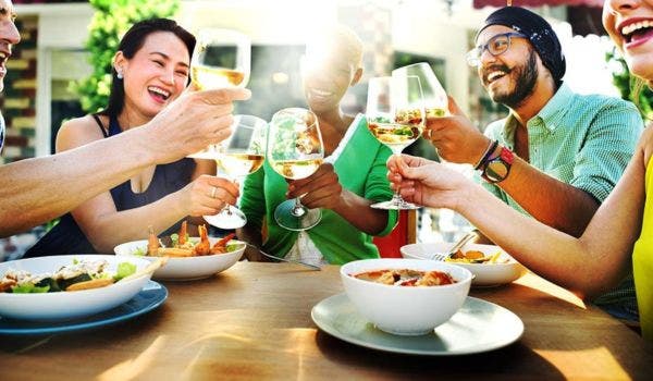 Diverse group of friends toasting with white wine glasses while dining outdoors, sharing a joyful meal together.