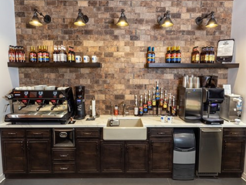 The back bar of a coffee shop with coffee makers and syrups on the countertop and shelves. A sink is in the middle of the counter and a Scotsman undercounter ice machine is pictured on the right side of the counter.
