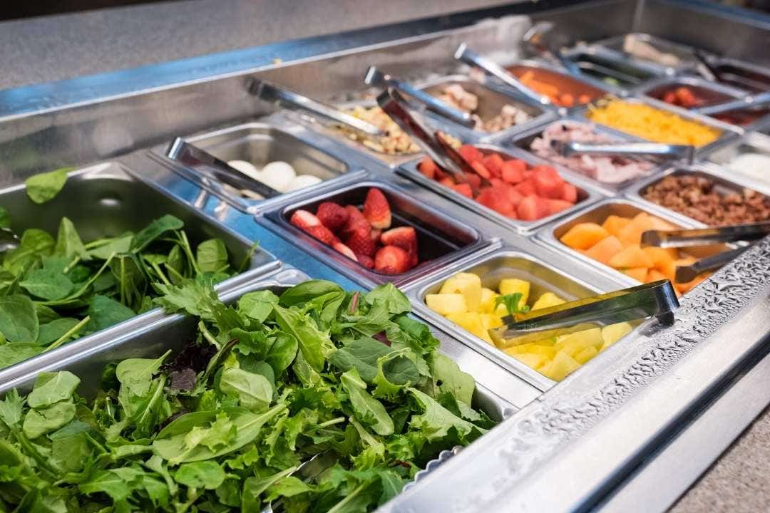 Fresh salad bar station with chopped greens and fruits in stainless steel pans
