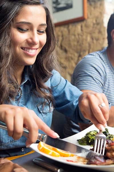 A woman uses a knife and fork to cut a piece of meat on a white plate while at a restaurant with a man seated next to her.